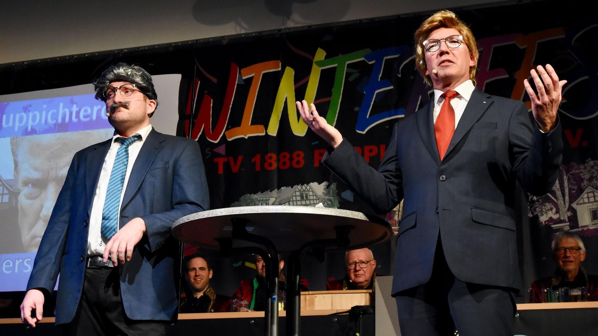 Zwei Männer auf einer Bühne im Karneval; der Mann rechts ist durch Anzug und Perücke als Donald trump zu erkennen.