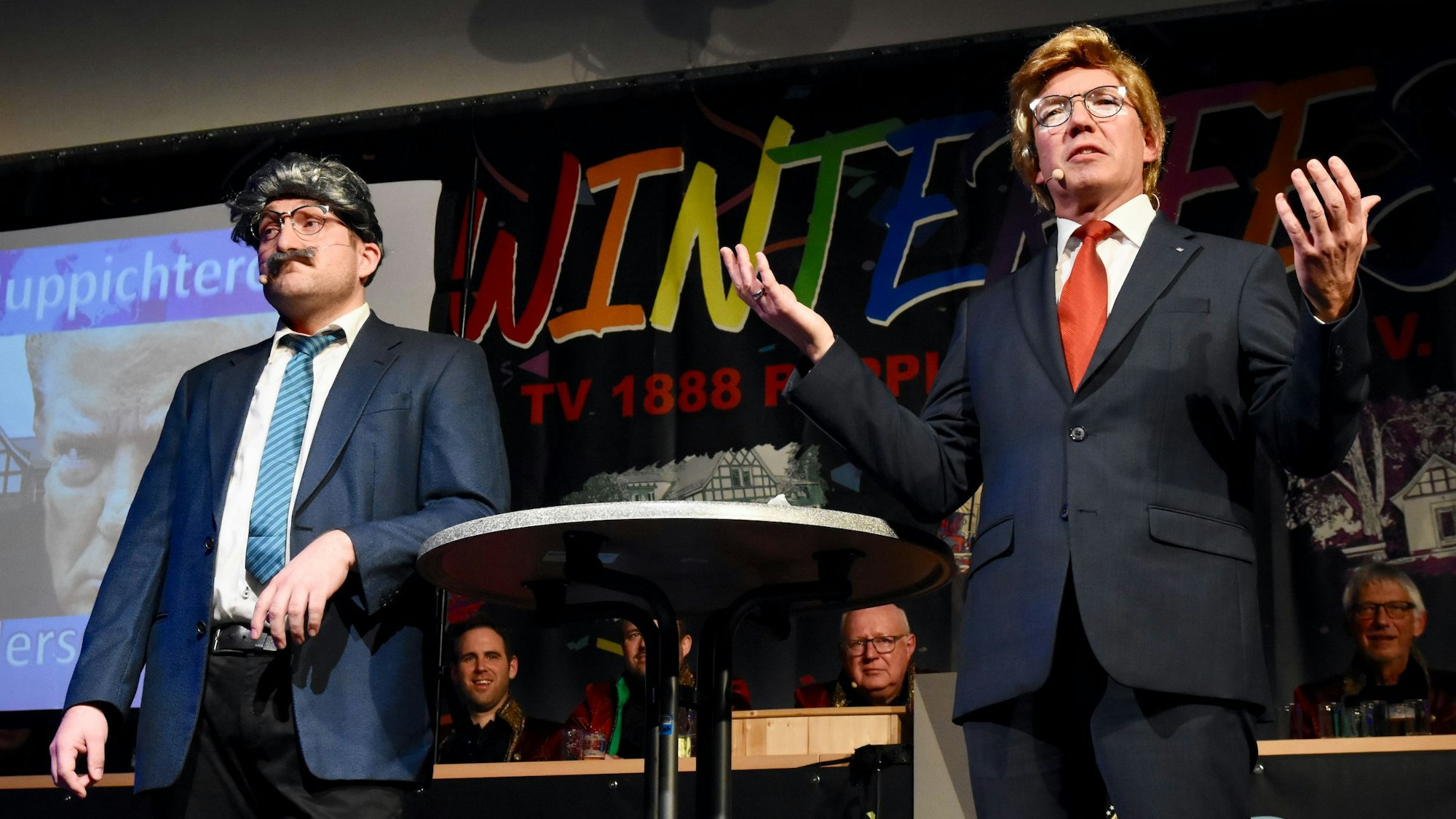 Zwei Männer auf einer Bühne im Karneval; der Mann rechts ist durch Anzug und Perücke als Donald trump zu erkennen.