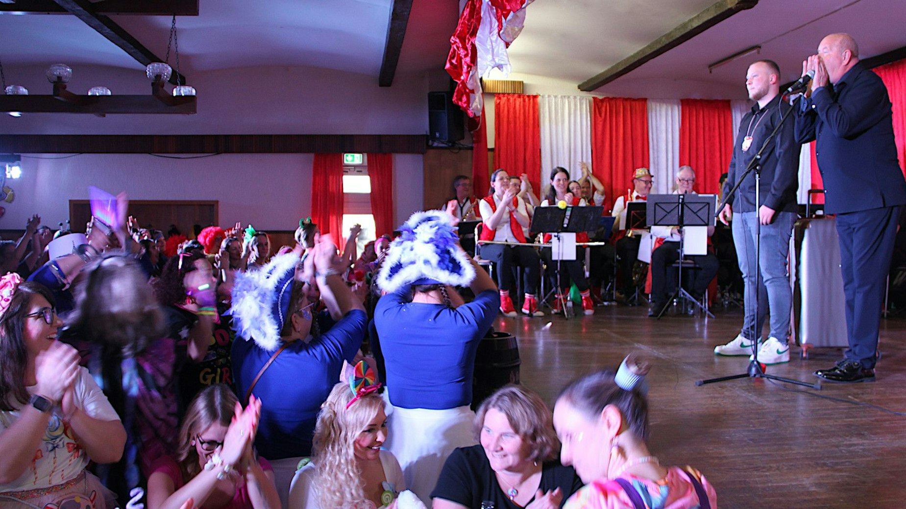 Auf einer Karnevalsbühne stehen zwei Männer, die bei der Mädchesitzung in Ripsdorf auftreten. Das Publikum applaudiert.