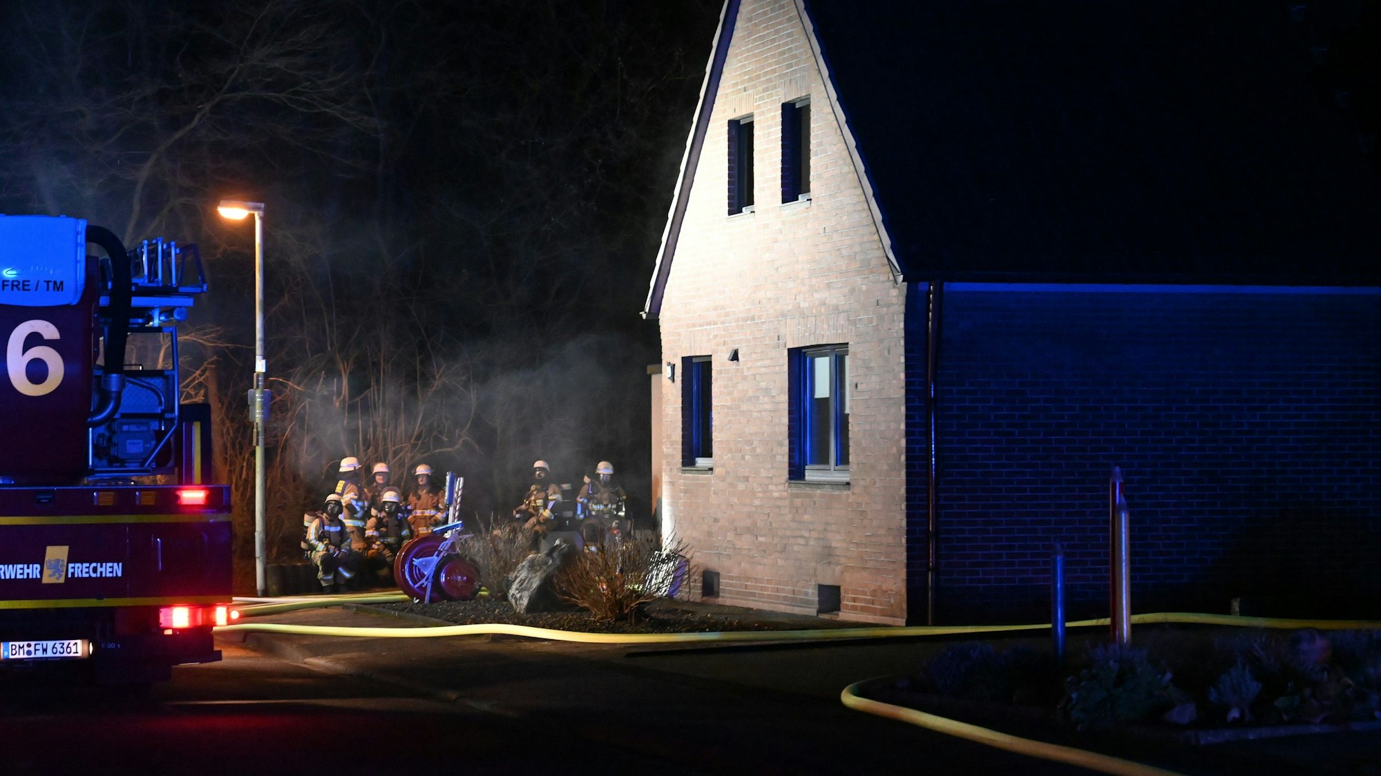 Feuerwehrleute, ein Einsatzfahrzeug und ein gelber dicker Schlauch sind vor einem Einfamilien