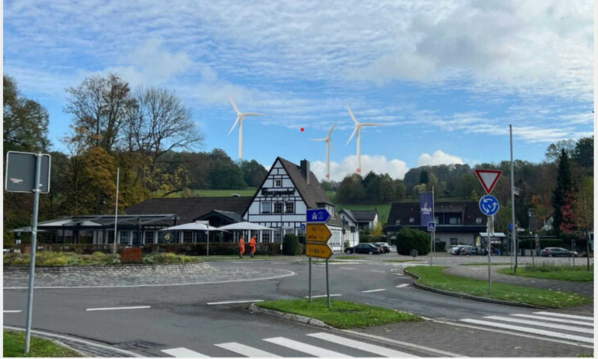 Fotocollage: Ein Kreisverkehr, dahinter ein Fachwerkhaus, am Horizont drei Windräder.
