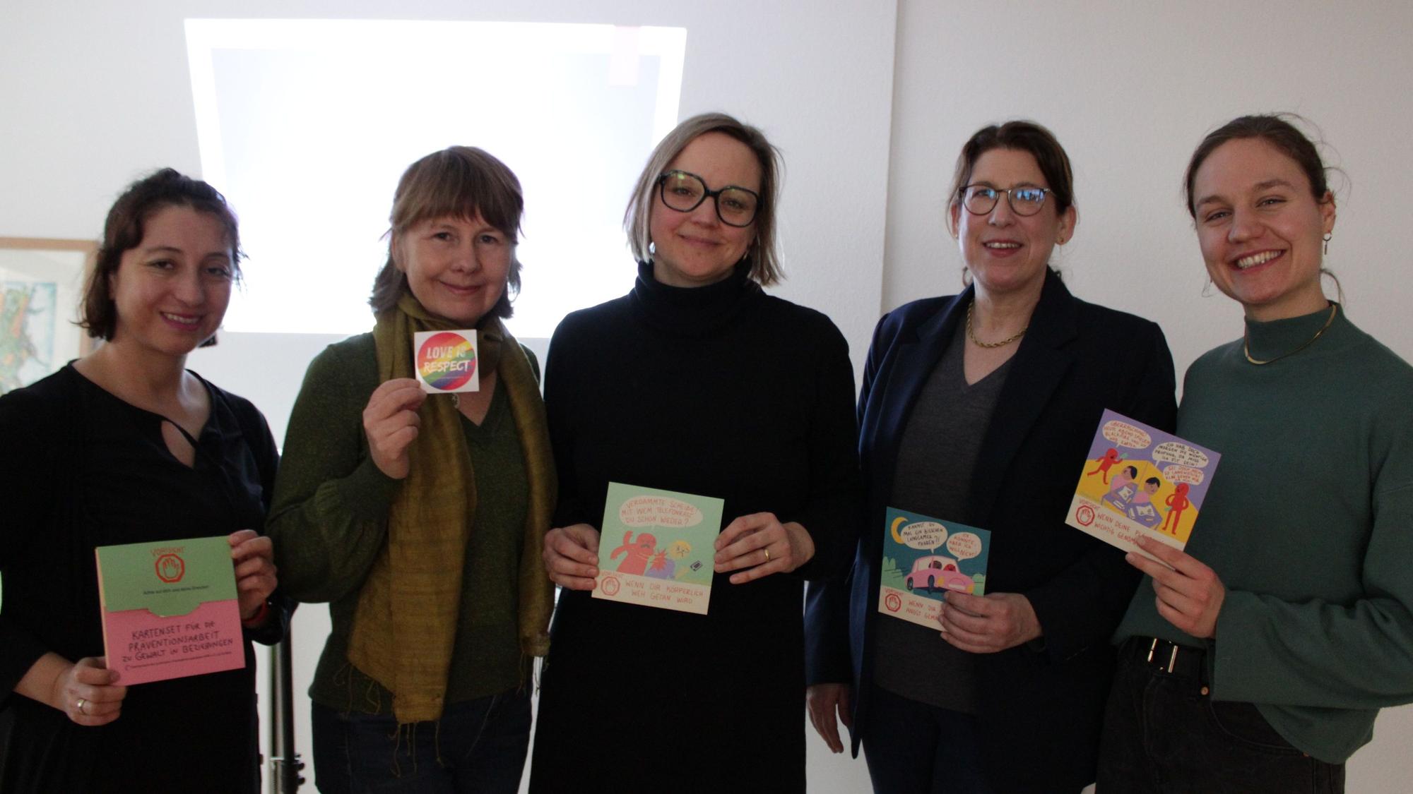 Fünf Frauen lächeln in die Kamera und halten Informationskarten aus dem Workshop „Liebe ist...“ in den Händen.