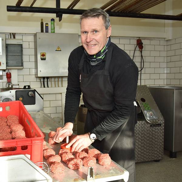 Ein Mann steht in schwarzer Arbeitsmontur an einem Tisch auf dem rote Lebensmittel-Boxen stehen und formt aus Hackfleisch Frikadellen.