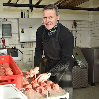 Ein Mann steht in schwarzer Arbeitsmontur an einem Tisch auf dem rote Lebensmittel-Boxen stehen und formt aus Hackfleisch Frikadellen.