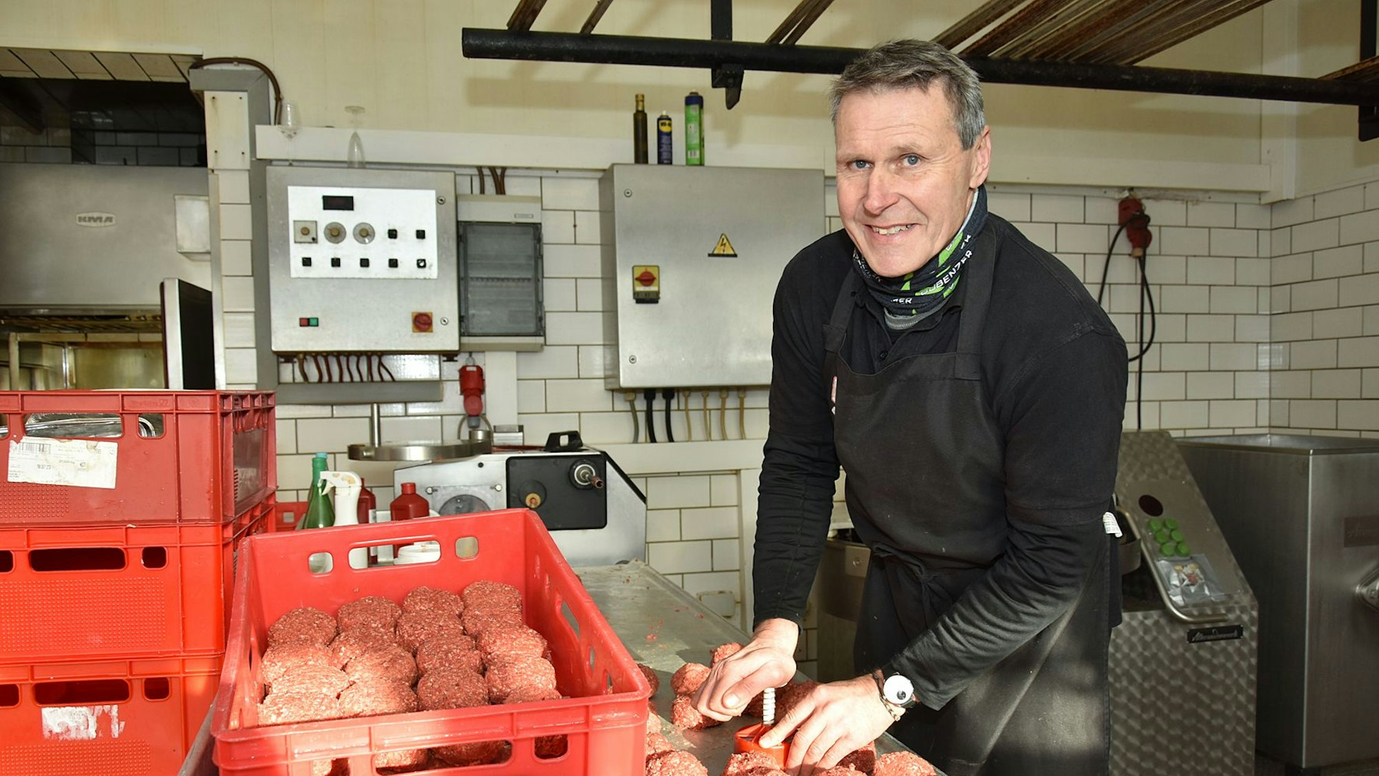 Ein Mann steht in schwarzer Arbeitsmontur an einem Tisch auf dem rote Lebensmittel-Boxen stehen und formt aus Hackfleisch Frikadellen.
