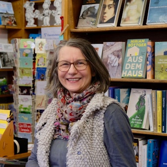 Eine Frau mit Brille, schulterlangem Haar und buntem Schal um den Hals steht lachend vor einem Bücherregal.