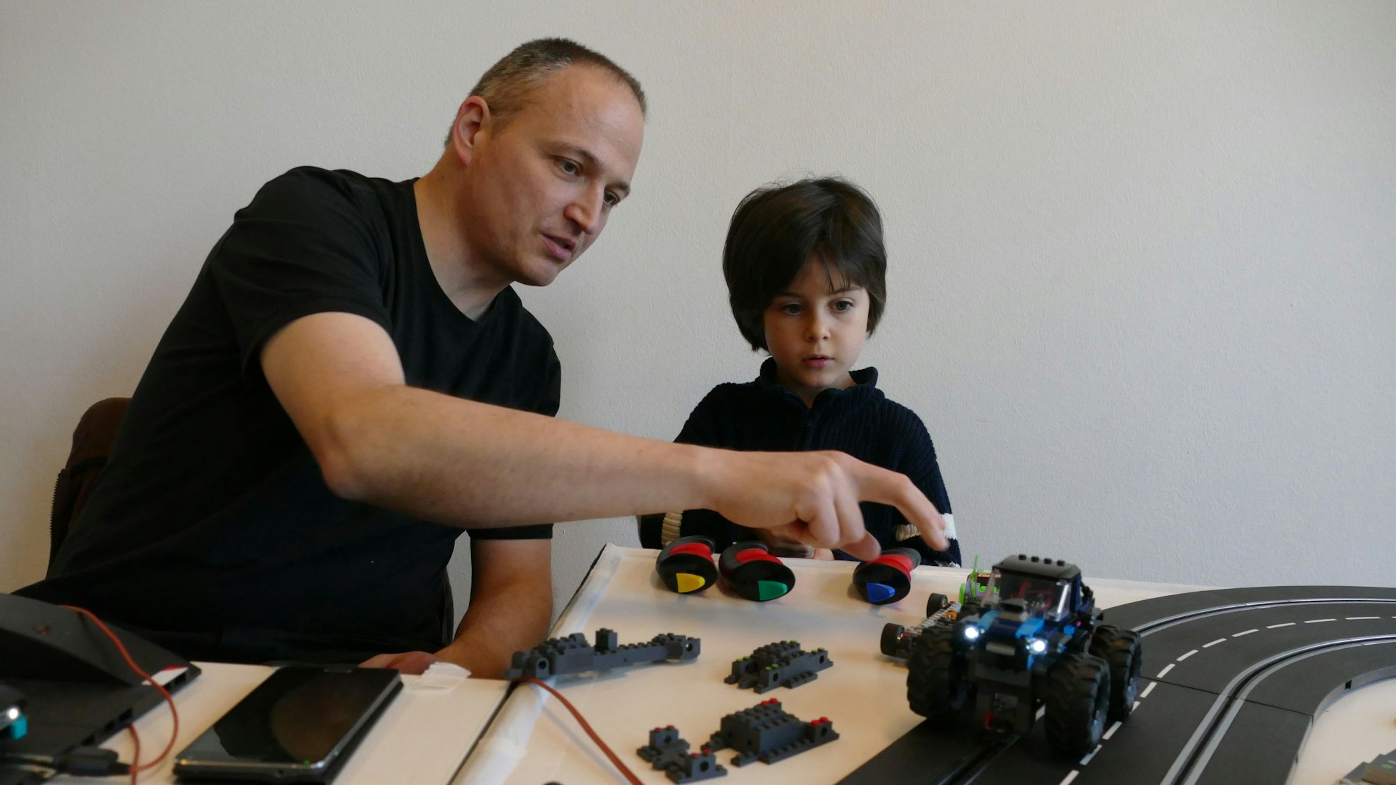 Ein Mann im schwarzen T-Shirt und ein Kind sitzen neben einer Spielzeugrennbahn.