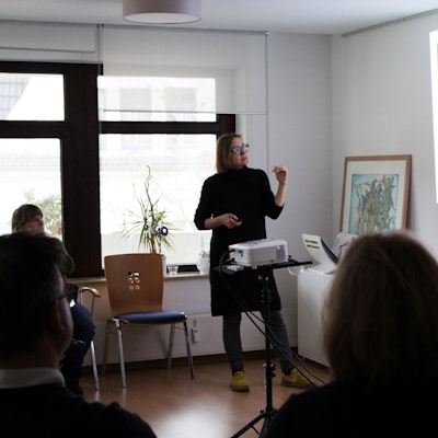 Eine Frau steht vor einem Beamer und deutet auf einen Comic. Eine Person darin fragt „ist das mein Handy?“, eine rot gefärbte Figur, die ein Handy in der Hand hält, sagt „Hast du etwa was zu verbergen?“