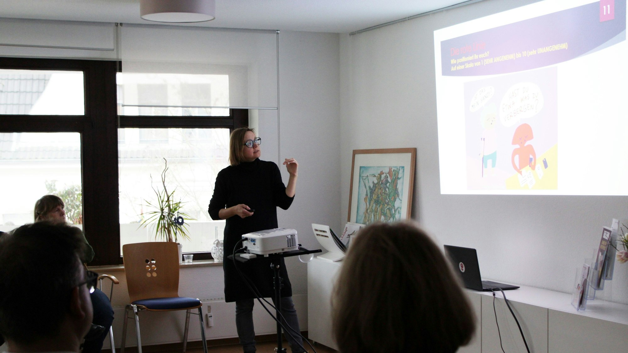 Eine Frau steht vor einem Beamer und deutet auf einen Comic. Eine Person darin fragt „ist das mein Handy?“, eine rot gefärbte Figur, die ein Handy in der Hand hält, sagt „Hast du etwa was zu verbergen?“