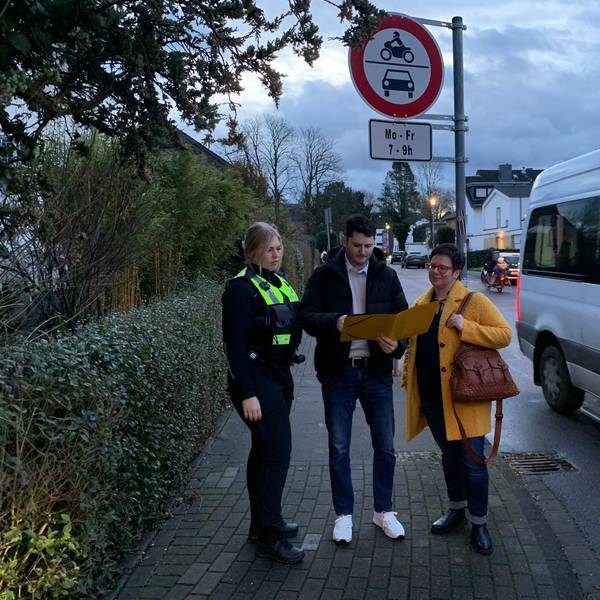 Drei Personen stehen an der Einmündung einer Straße und schauen in eine aufgeschlagene Mappe.