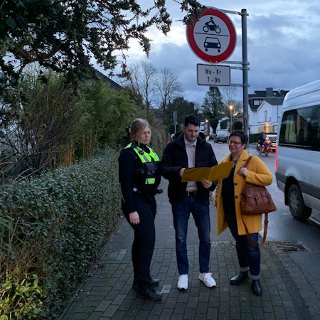 Drei Personen stehen an der Einmündung einer Straße und schauen in eine aufgeschlagene Mappe.
