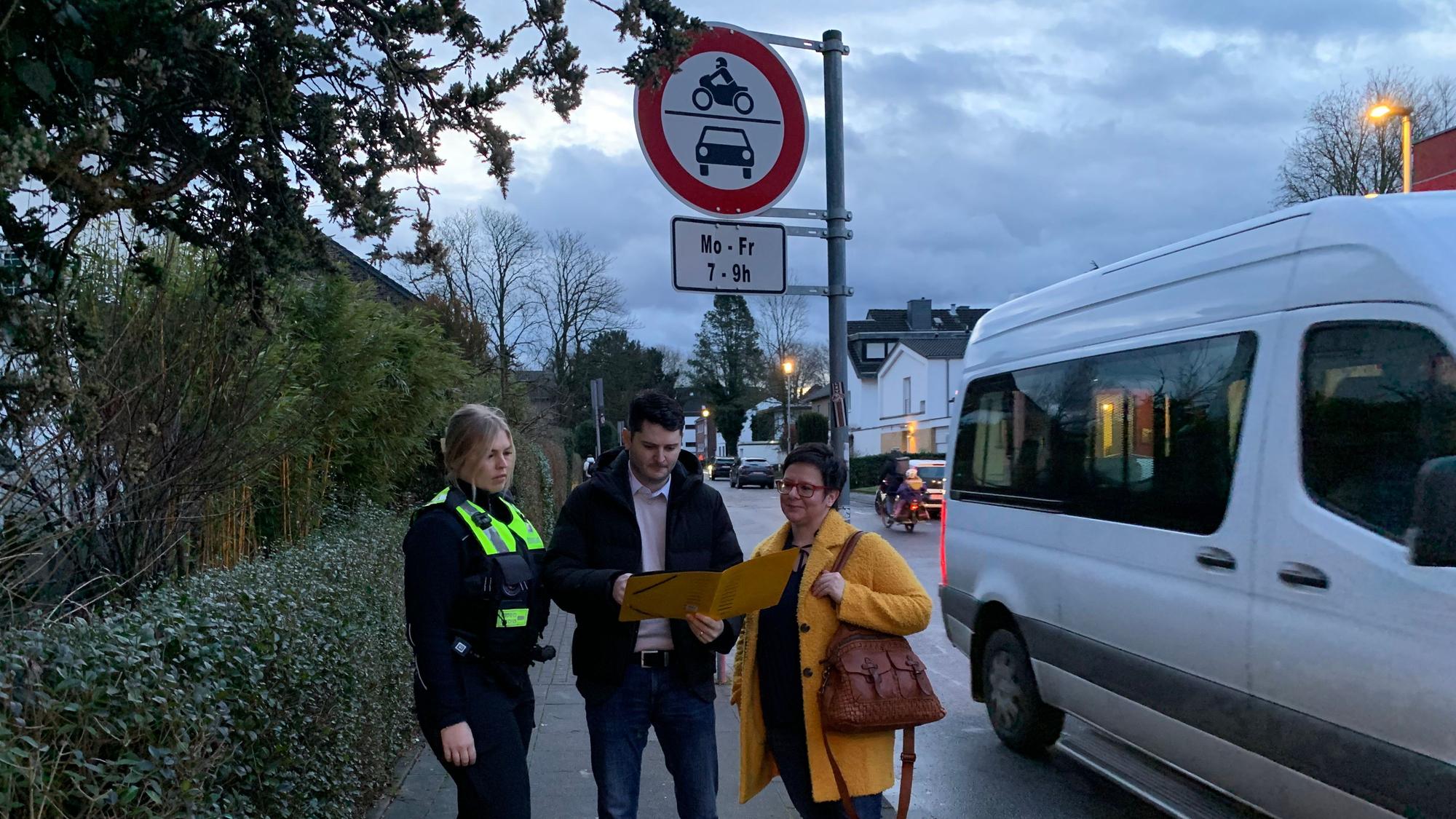 Drei Personen stehen an der Einmündung einer Straße und schauen in eine aufgeschlagene Mappe.