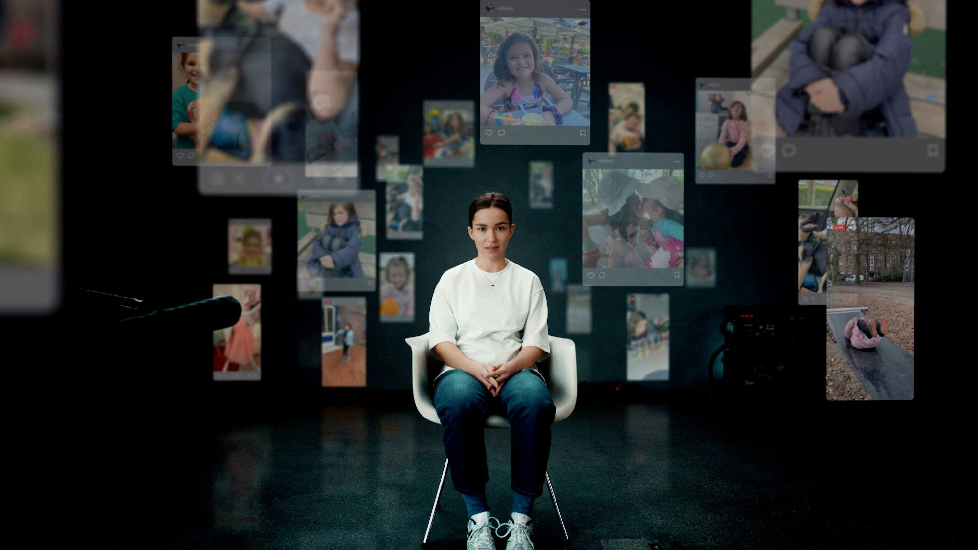 Eine junge Frau sitzt auf einem Stuhl, im Hintergrund ist eine Wand mit vielen Kinderbildern zu sehen.