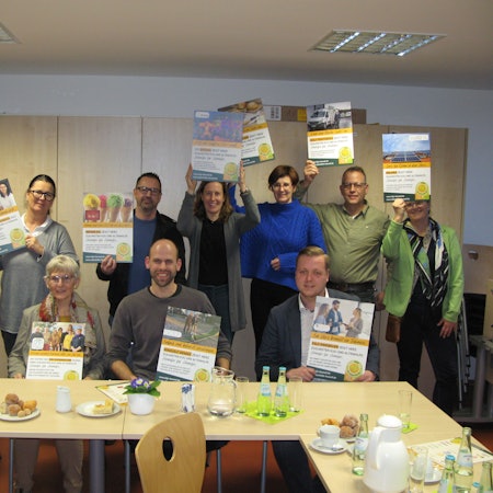 Zwei Frauen und zwei Männer sitzen nebeneinander an einem Tisch. Dahinter stehen vier Frauen und drei Männer. Alle halten Plakate hoch.
