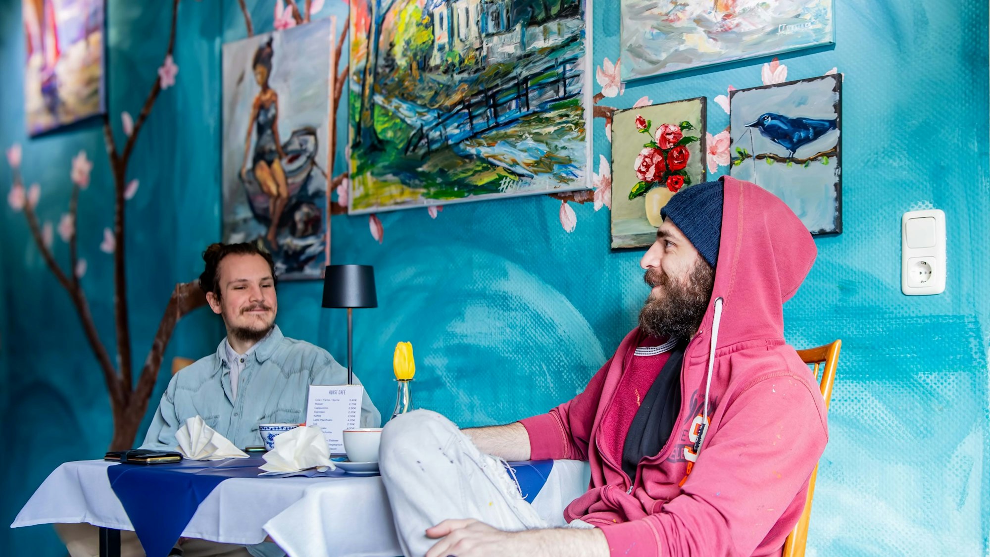Das Bild zeigt zwei Männer an einem Tisch im Café sitzen. Im Hintergrund hängen Bilder an der Wand.