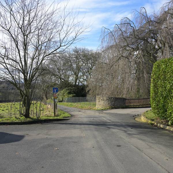 Eine Straße endet als Feldweg. Rechts eine hohe, grüne Laubhecke, dahinter eine Grundstückseinfahrt.