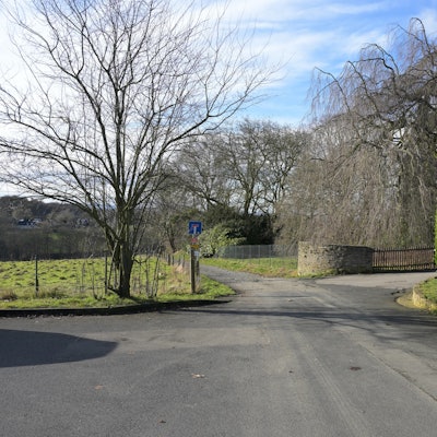 Eine Straße endet als Feldweg. Rechts eine hohe, grüne Laubhecke, dahinter eine Grundstückseinfahrt.