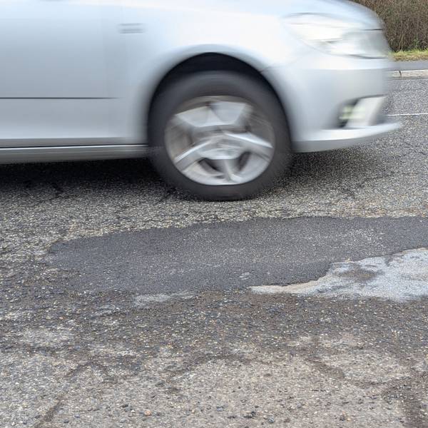 Das Foto zeigt das reparierte Schlagloch im Asphalt der Luxemburger Straße, dahinter ein vorbeifahrendes Auto.