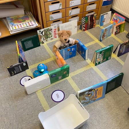 Ein mit Bücher aufgebauter Parcours auf einem aufgeklebten Gitternetz mit einem blauen, aus vier Kugeln bestehenden Roboter