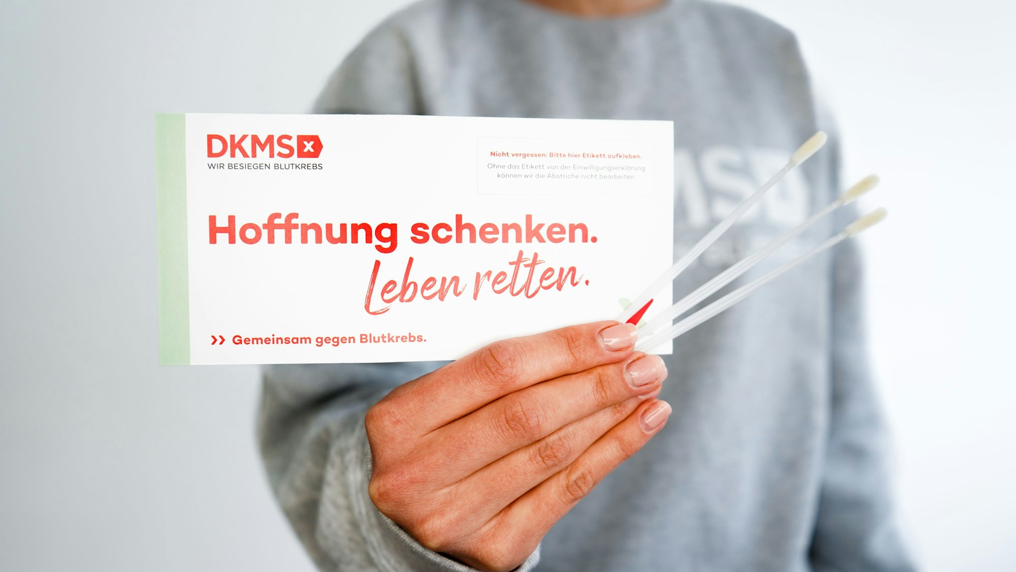 Das Foto zeigt eine Hand mit drei Wattestäbchen und einem Testset der DKMS.
