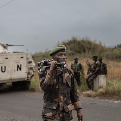 Ein Soldat mit Beret und geschultertem Gewehr vor einem hellen Panzer mit der Aufschrift „UN“.