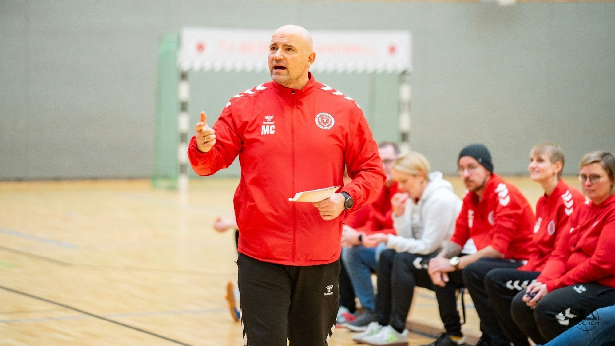 Das Bild zeigt einen Handballtrainer in einer roten Jacke vor seiner Bank