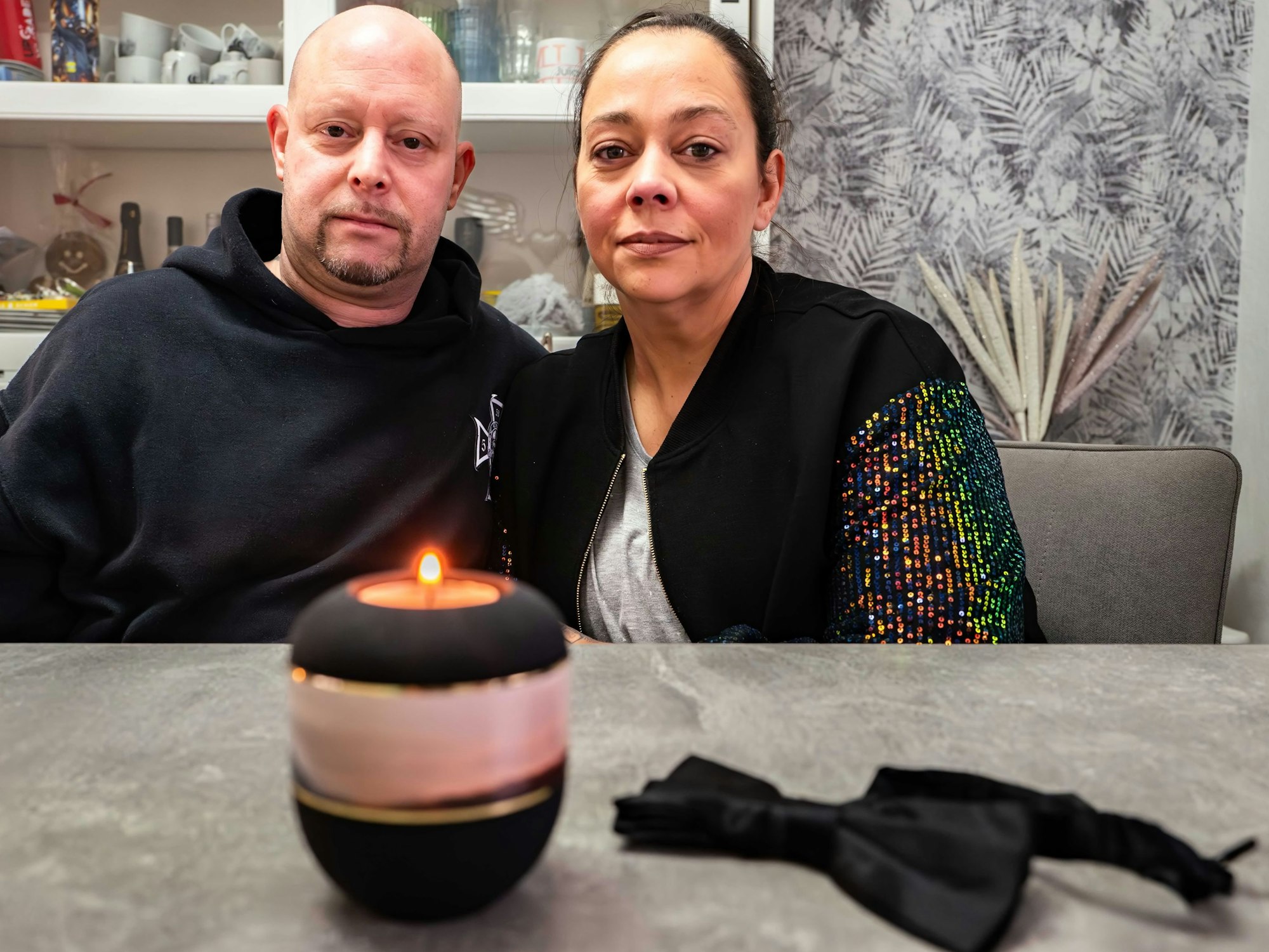 Jasmin Glasmacher und ihr Mann Markus sitzen an einem Tisch. Darauf steht eine Kerze, neben dieser liegt eine schwarze Fliege.