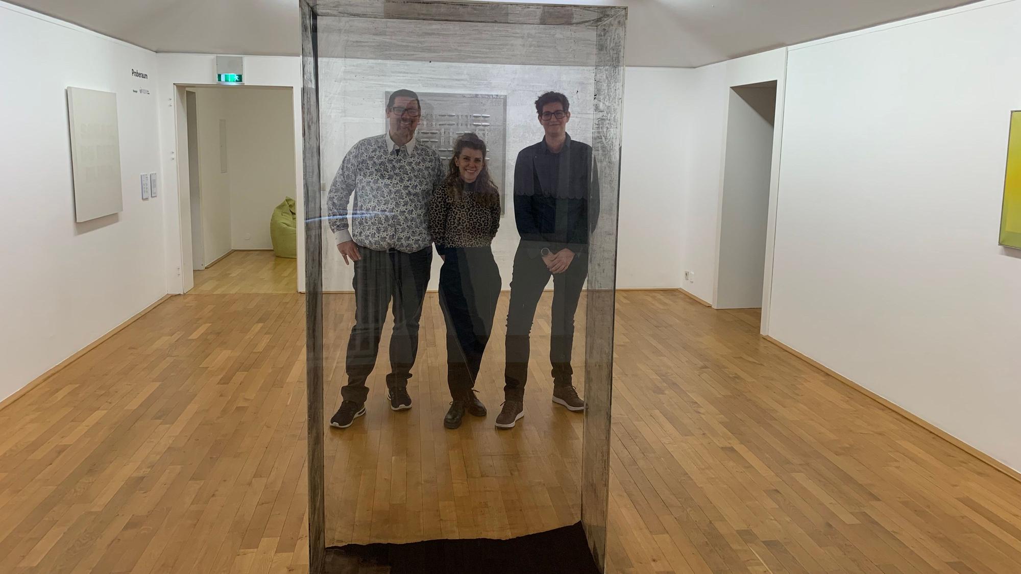 Drei Personen stehen hinter einem Kasten mit durchsichtigen, aber dreckigen Scheiben in einem Ausstellungsraum.