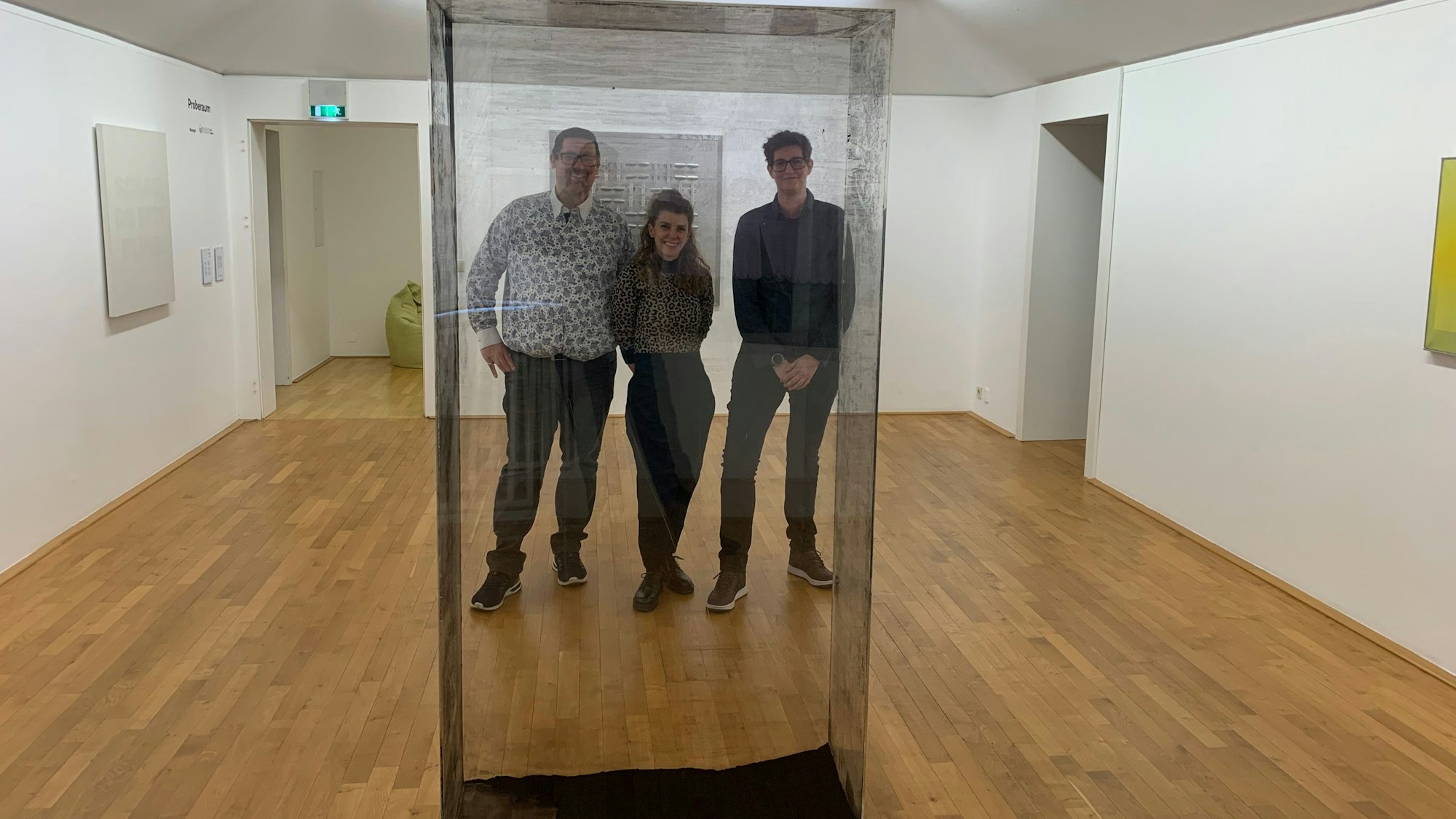 Drei Personen stehen hinter einem Kasten mit durchsichtigen, aber dreckigen Scheiben in einem Ausstellungsraum.
