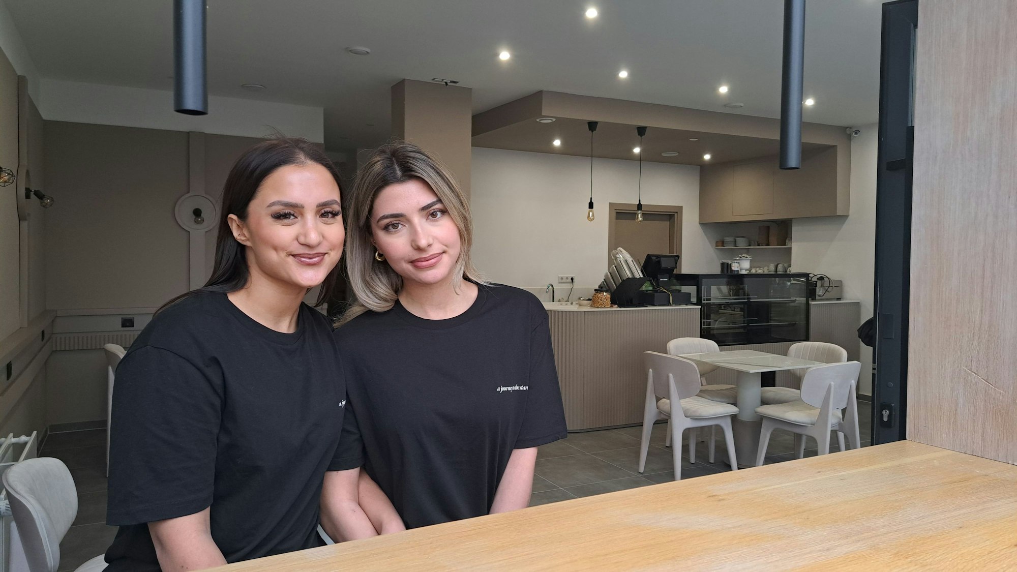 Zwei Frauen mit langen Haaren tragen schwarze T-Shirts und sitzen an der Holztheke ihres Cafés.