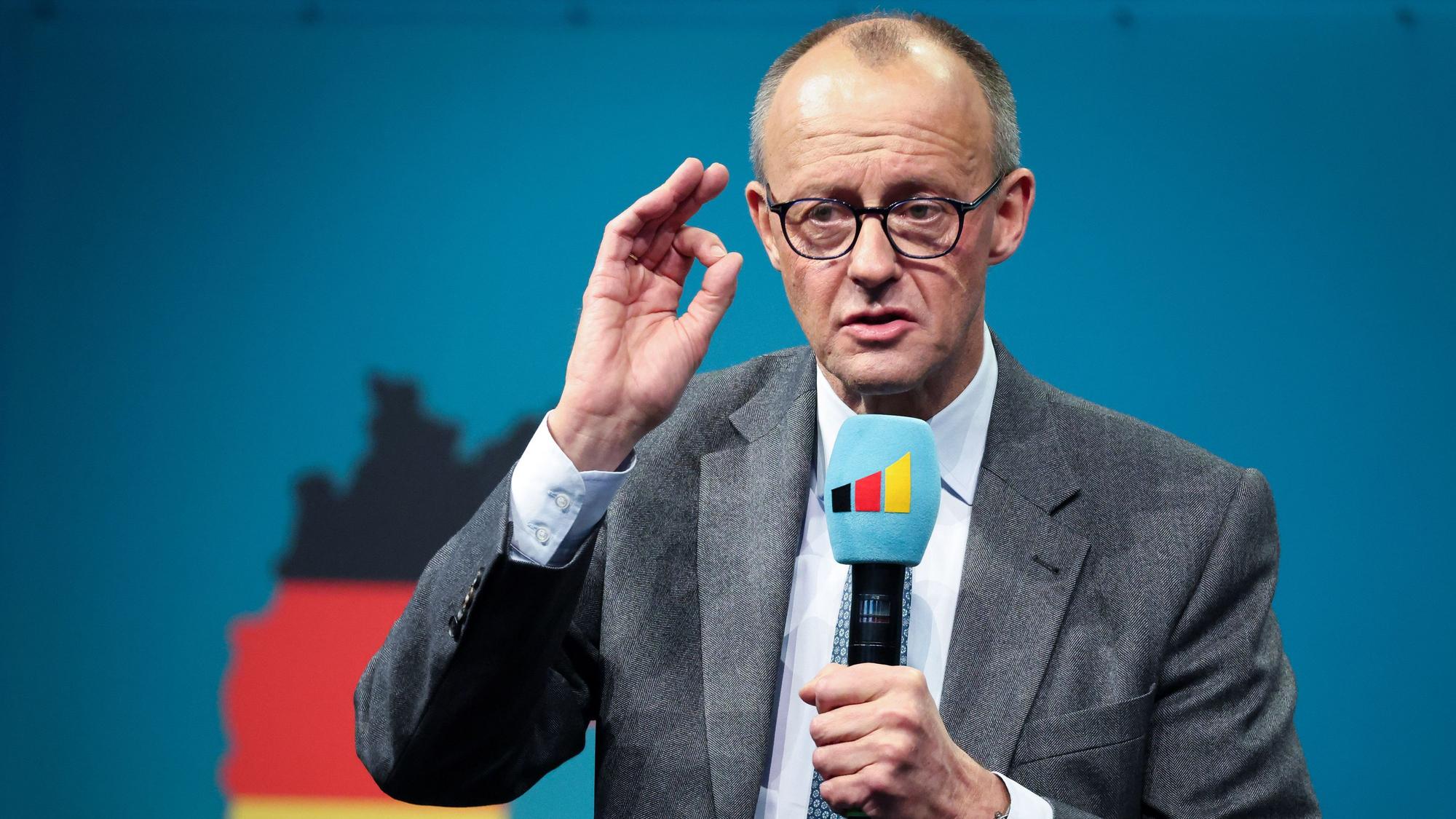 CDU-Kanzlerkandidat Friedrich Merz während einer Wahlkampfveranstaltung. Er trägt einen grauen Anzug und hält ein Mikrofon in der linken Hand, während er seine Worte gestisch mit der rechten, erhobenen Hand unterstreicht. Er steht vor einem blauen Hintergrund, auf dem undeutlich die Umrisse von Deutschland in den Farben schwarz-rot-gold zu sehen sind.