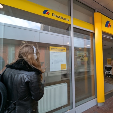 Das Foto zeigt eine Passante vor einem Aushang im Schaufenster der Postbank-Filiale im Hürth-Park.