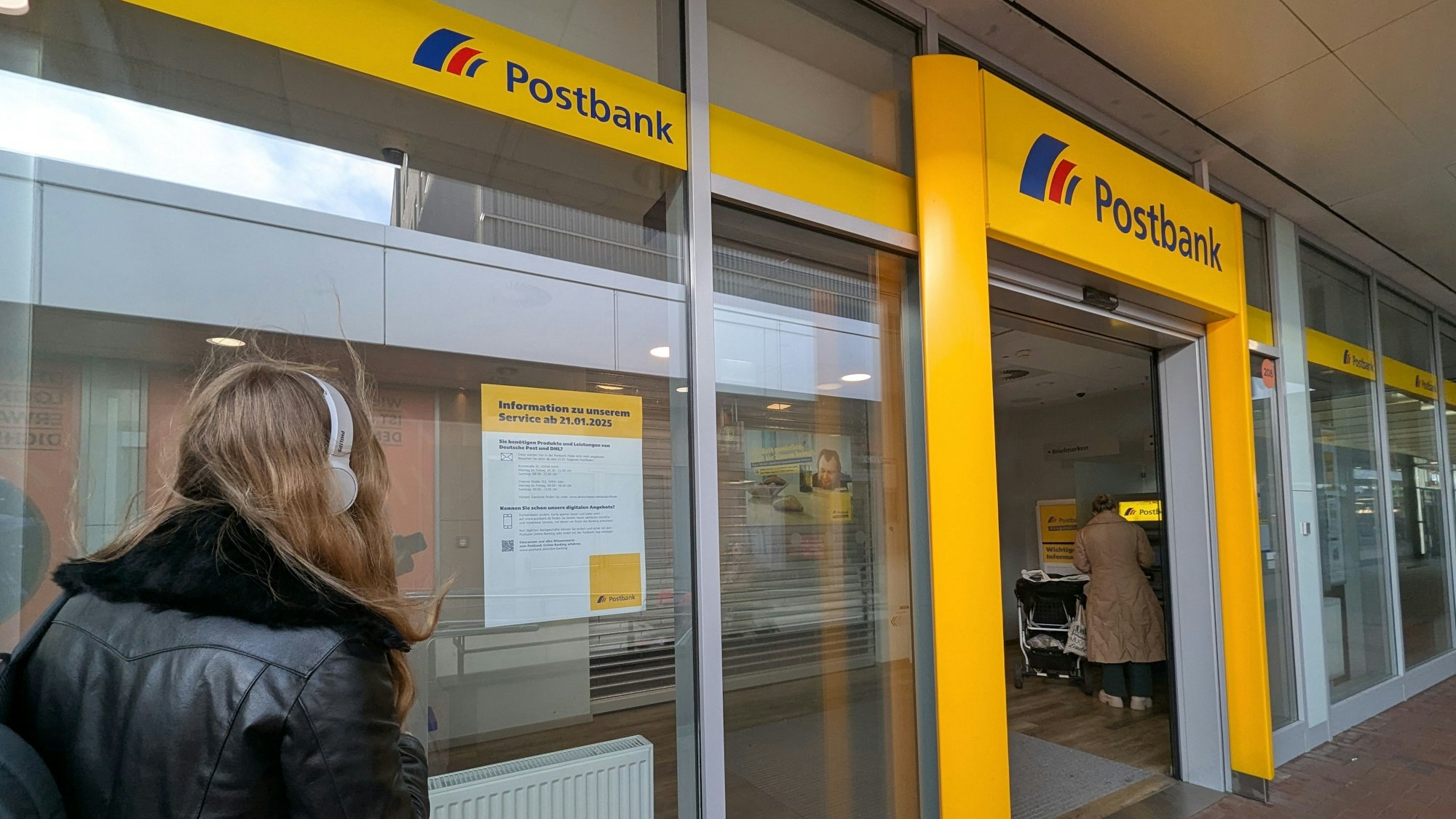 Das Foto zeigt eine Passante vor einem Aushang im Schaufenster der Postbank-Filiale im Hürth-Park.