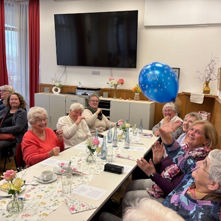 Eine Gruppe älterer Frauen, an einem gedeckten Tisch, spielt sich einen Luftballon zu.