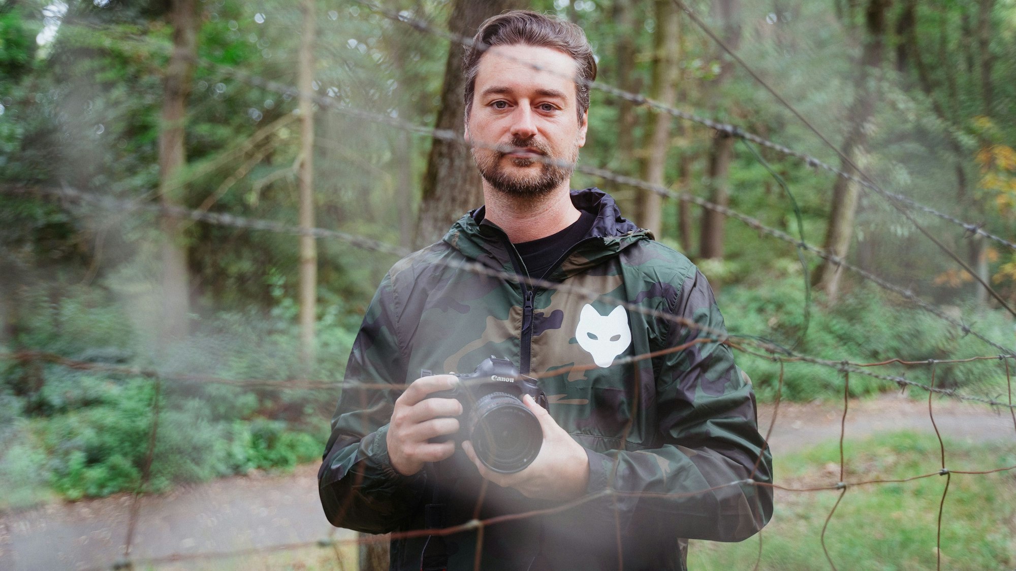 Ein Mann mit einer Kamera steht vor Bäumen und hinter einem Stacheldrahtzaun.