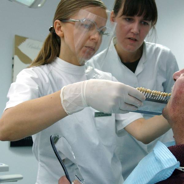 Zwei Frauen in medizinischer Kleidung stehen vor einem Mann und halten ihm Zahnprothesen an den Mund.