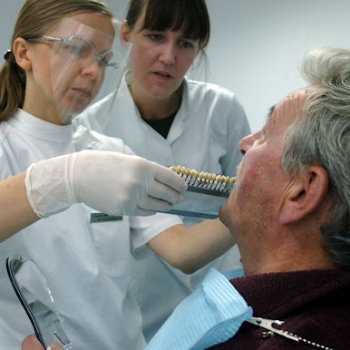 Zwei Frauen in medizinischer Kleidung stehen vor einem Mann und halten ihm Zahnprothesen an den Mund.