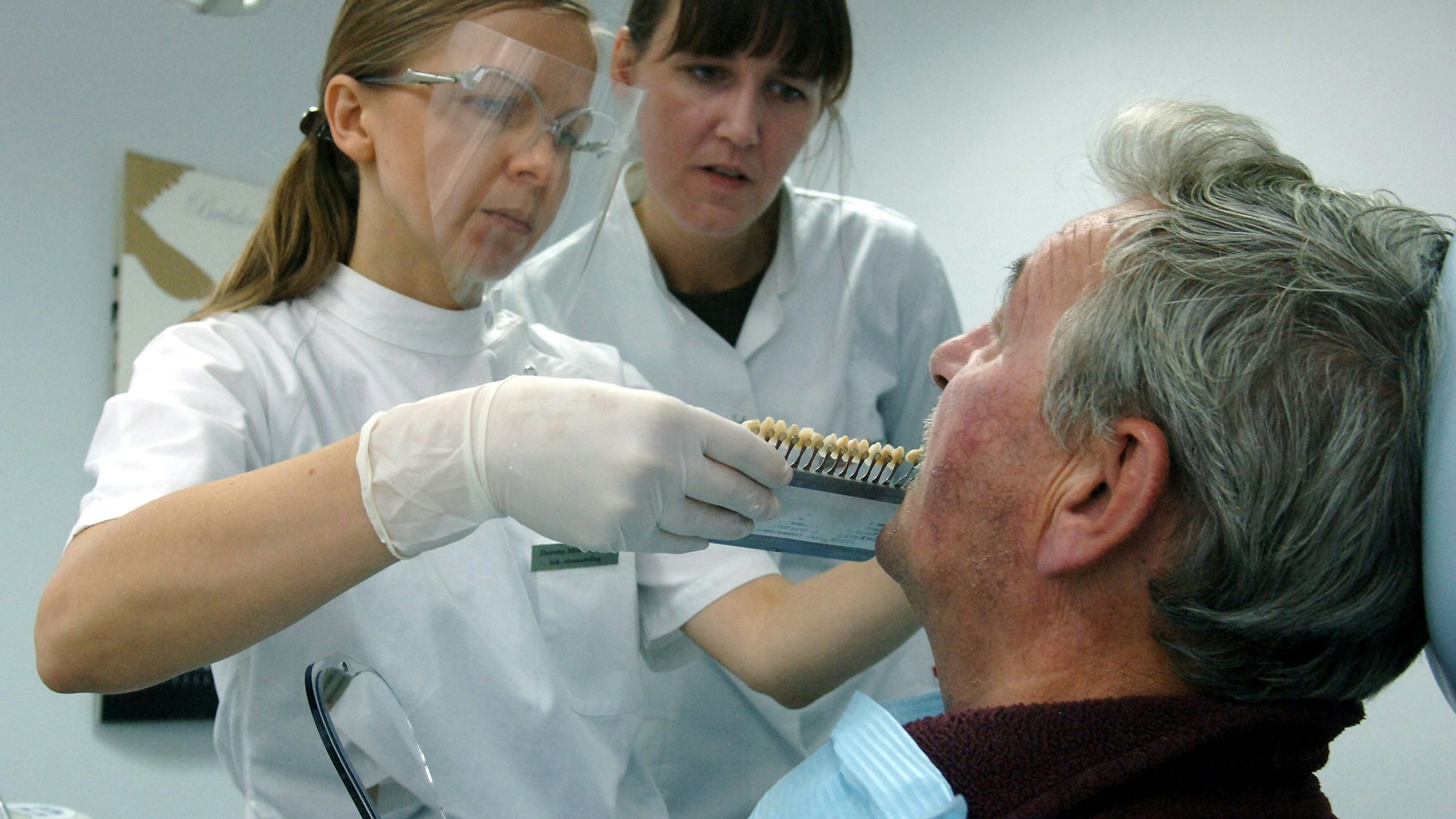 Zwei Frauen in medizinischer Kleidung stehen vor einem Mann und halten ihm Zahnprothesen an den Mund.