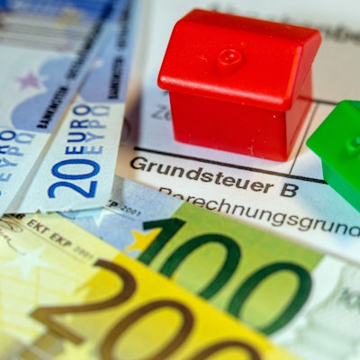 Ein Spielzeughaus und die Nachbildung von Euro-Geldscheinen liegen auf einem Abgabenbescheid für die Entrichtung der Grundsteuer.
