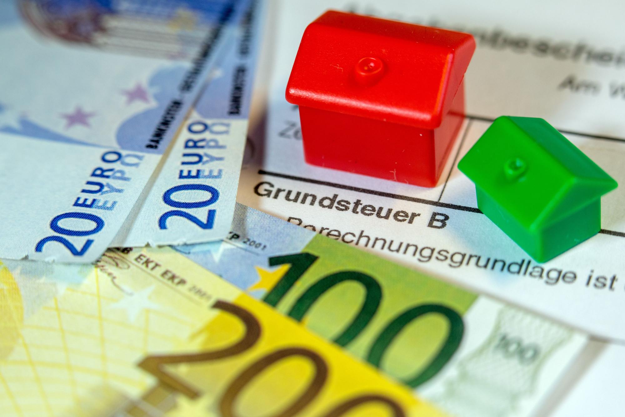 Ein Spielzeughaus und die Nachbildung von Euro-Geldscheinen liegen auf einem Abgabenbescheid für die Entrichtung der Grundsteuer. (Symbolbild)