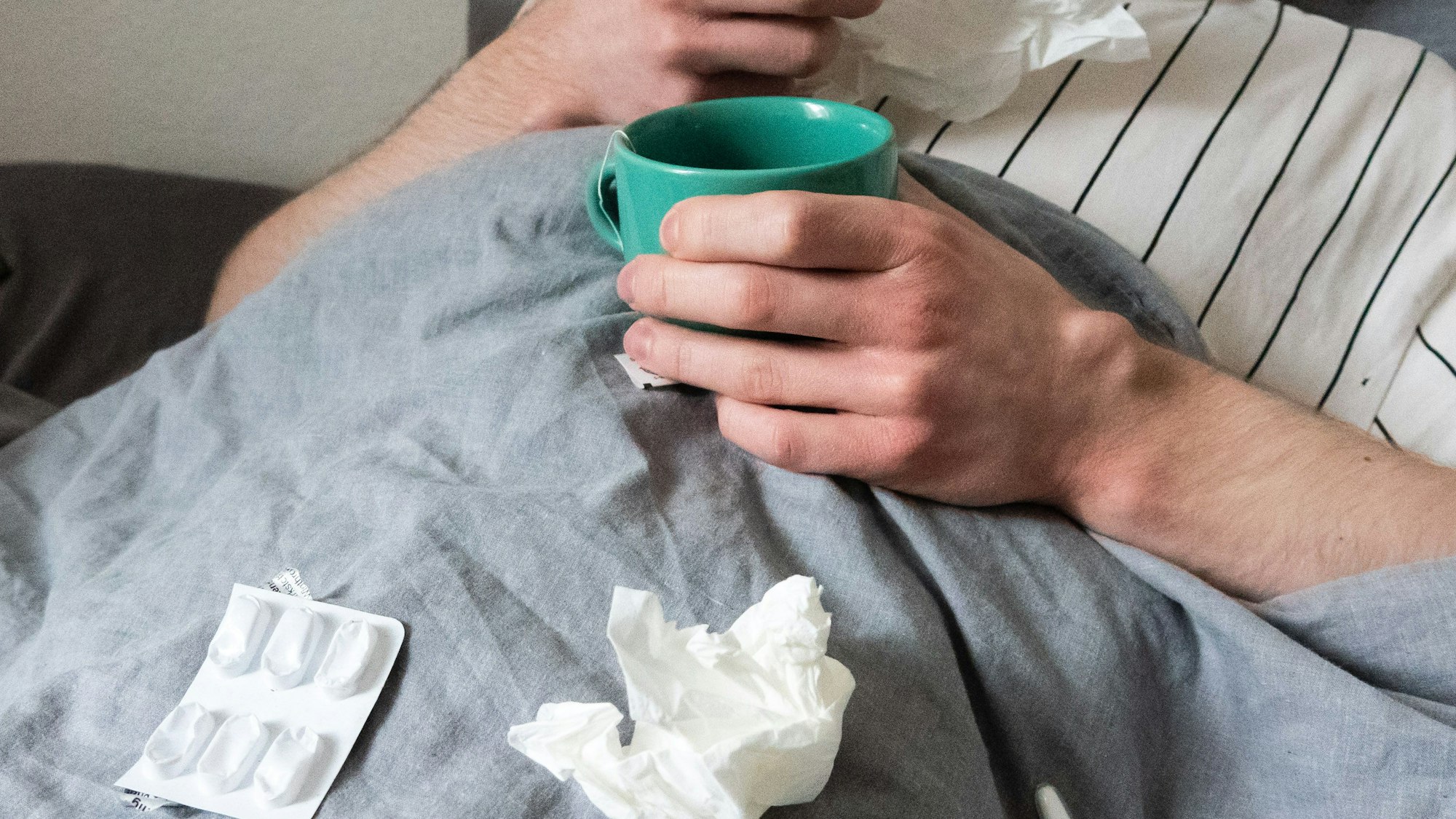 Ein Mann liegt mit Taschentüchern, Teetasse, Nasenspray und Tabletten im Bett (gestellte Szene, Archivbild)