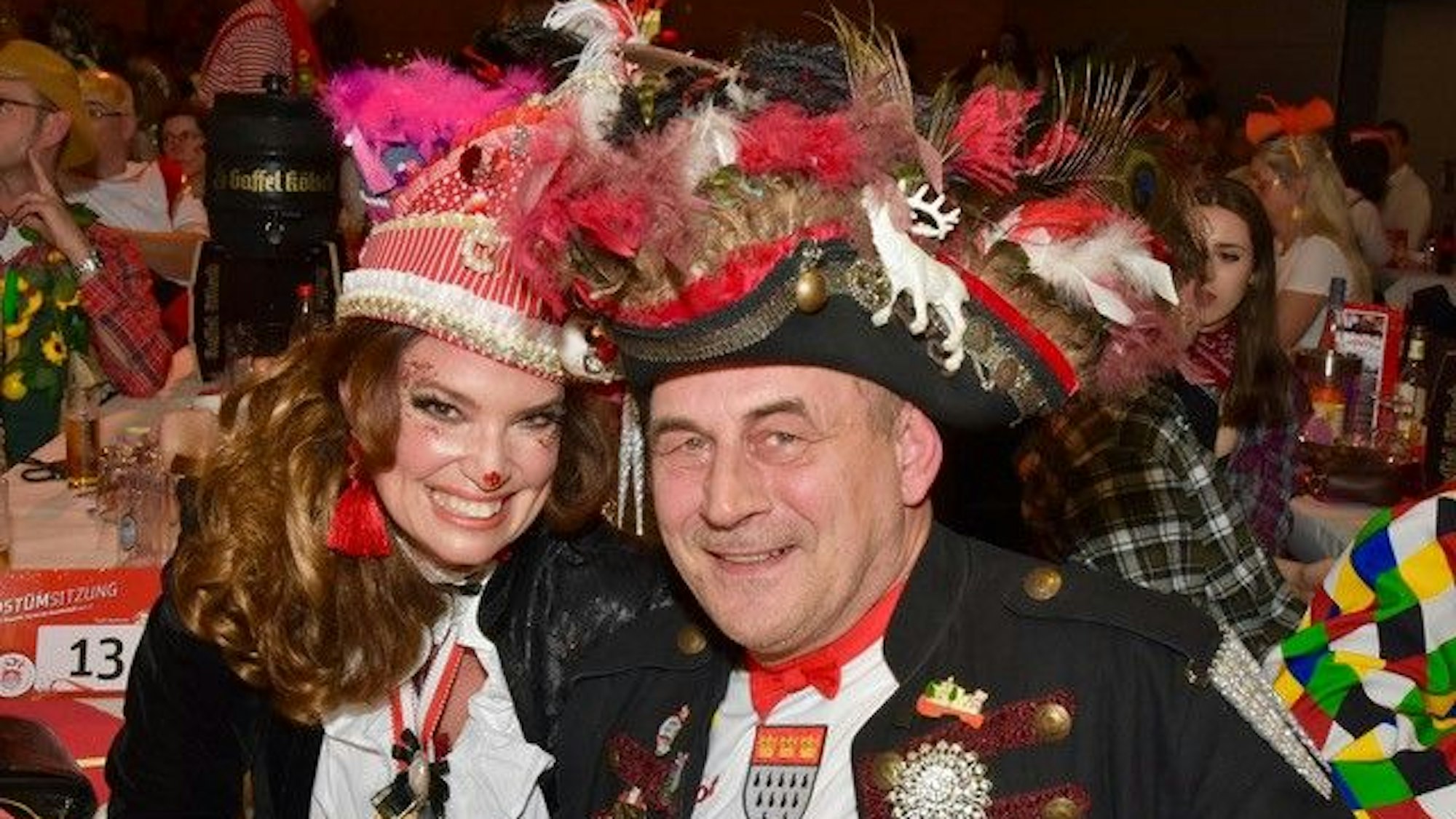 Ein Mann und eine Frau mit bunt geschmückten Hüten bei einer Karnevalssitzung.