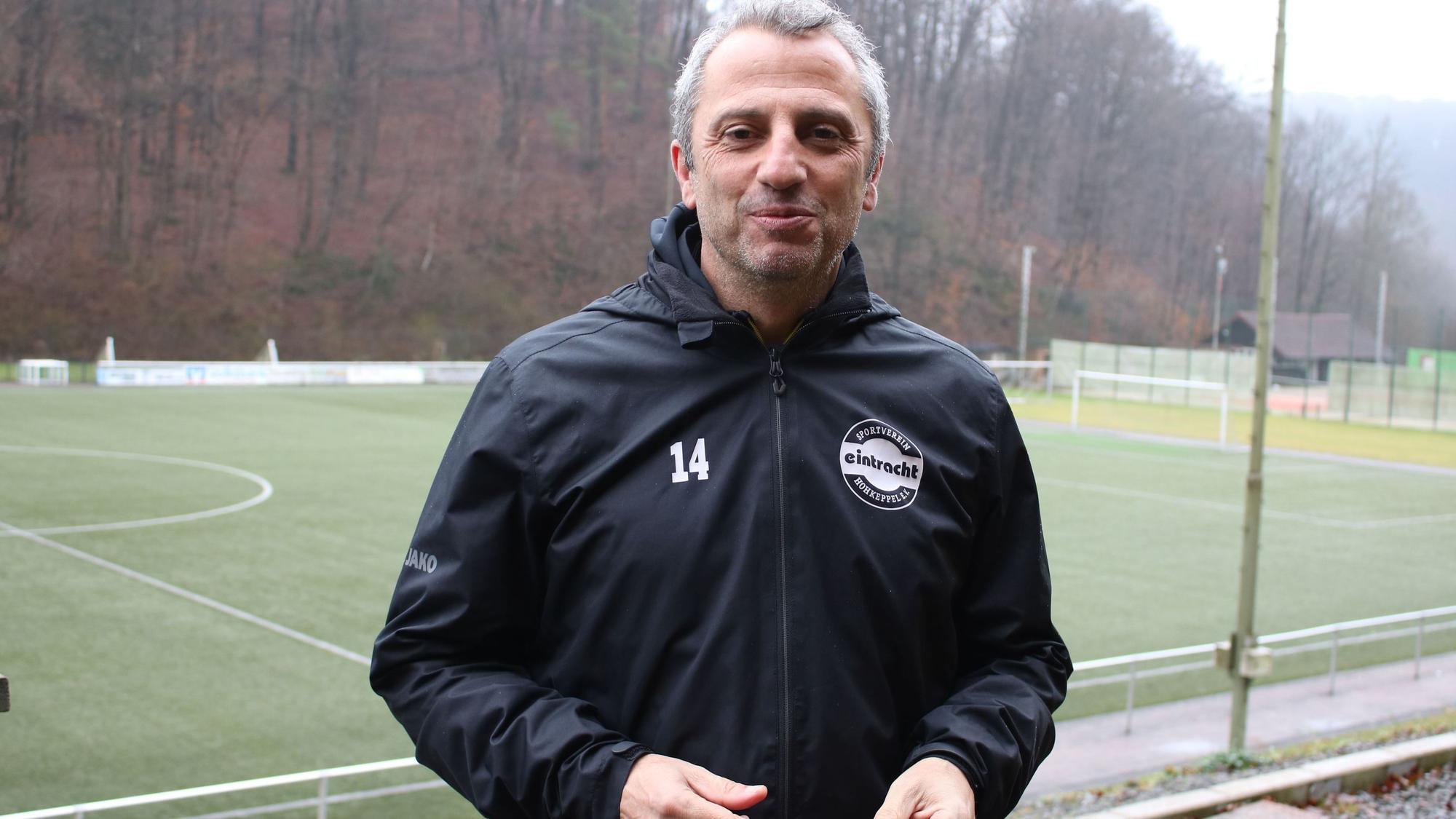 Ein Fußballtrainer steht auf einer Tribüne, im Hintergrund ist ein Fußballplatz zu sehen.