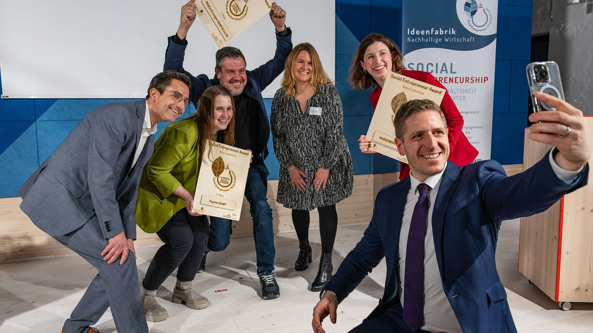 Im Vordergrund hat Landrat Markus Ramers ein Handy zum Fotografieren gezückt, während die drei Erstplatzierten juveld in die kamera schauen.