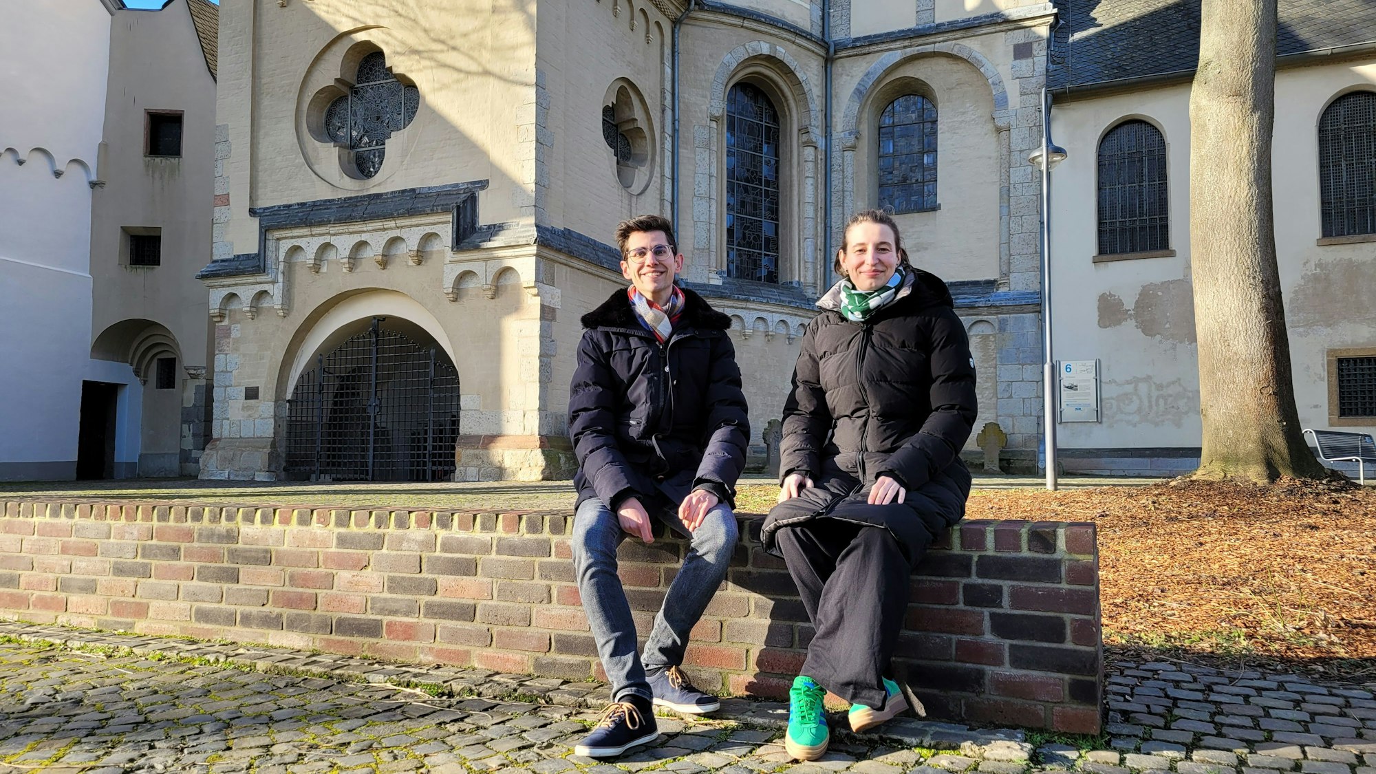 Ein Frau und ein Mann sitzen auf einer Mauer. Im Hintergrund die Abteikirche St. Nikolaus in Brauweiler.