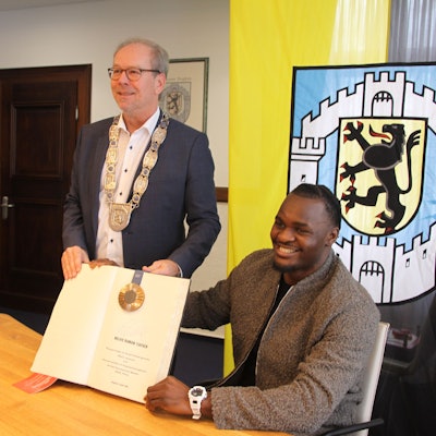 Das Bild zeigt den strahlenden Schwergewichtsboxer Nelvie Tiafack sitzend am Tisch und dahinter den Bürgermeister, beide halten das Goldene Buch in die Kamera.