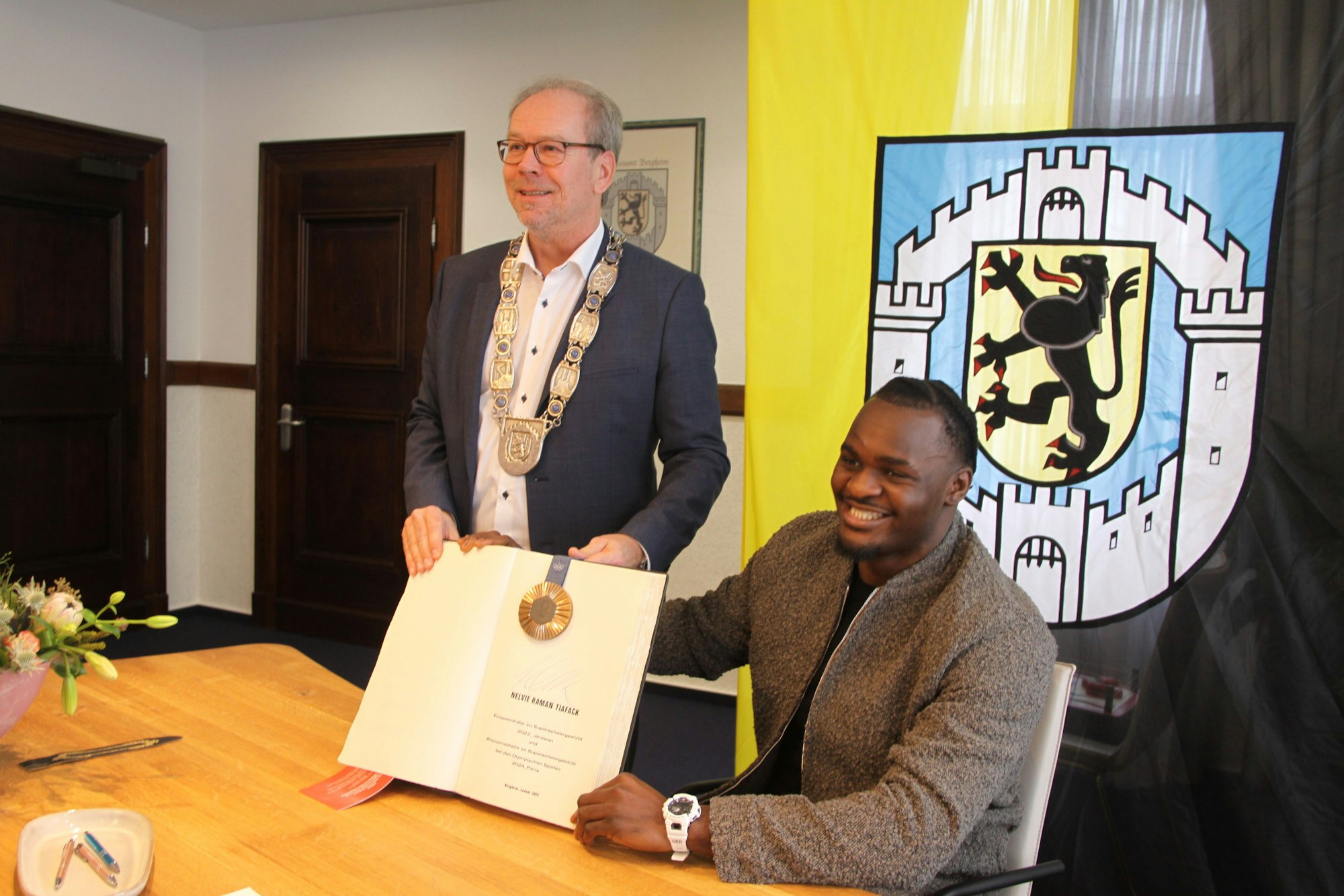 Das Bild zeigt den strahlenden Schwergewichtsboxer Nelvie Tiafack sitzend am Tisch und dahinter den Bürgermeister, beide halten das Goldene Buch in die Kamera.