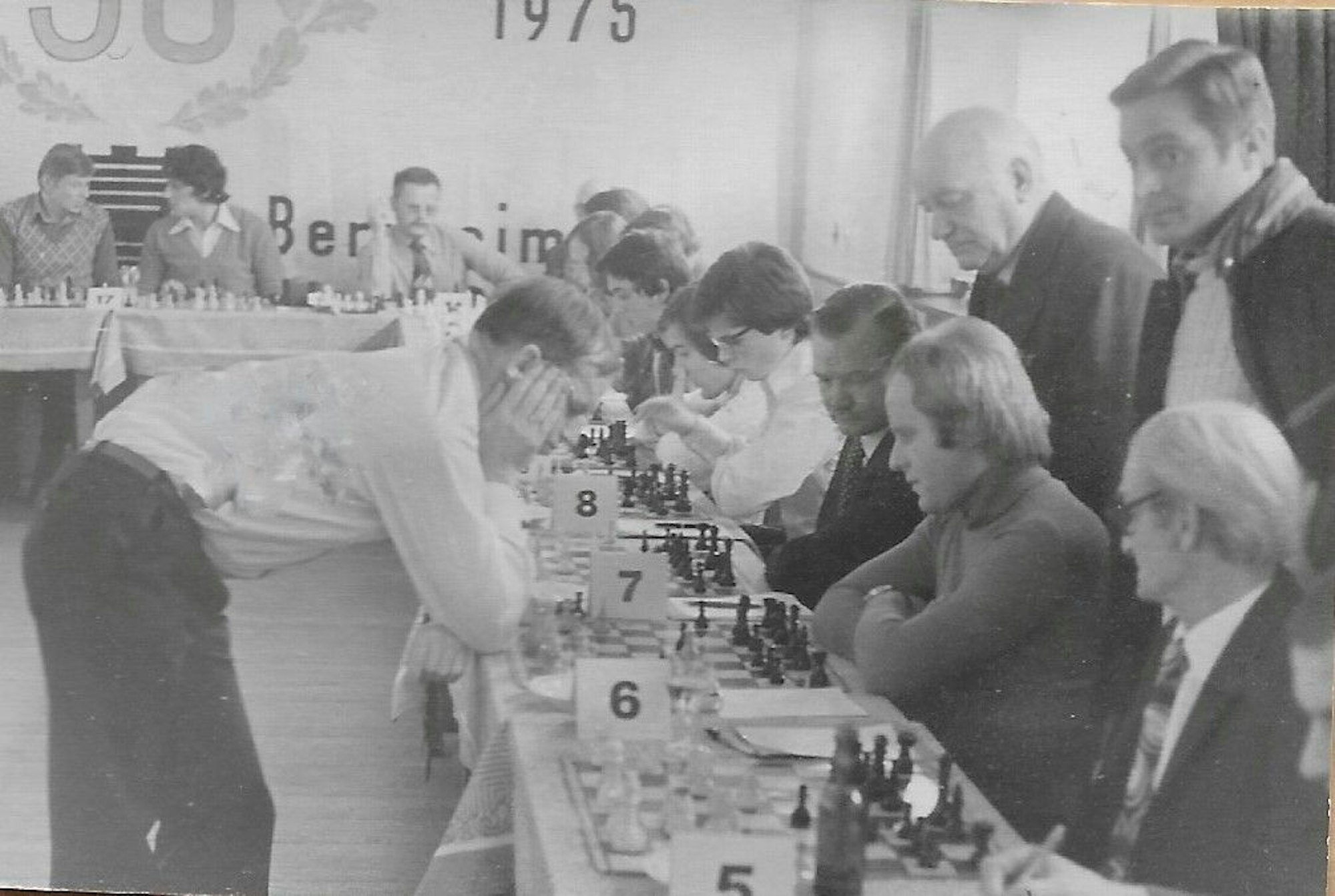 Eine ganze Tischreihe mit Schachbrettern ist zu sehen, an jedem Brett jeweils ein Gegner für Großmeister Wolfgang Unzicker. Der steht über einem der Bretter gebeugt und denkt über den nächsten Zug nach.