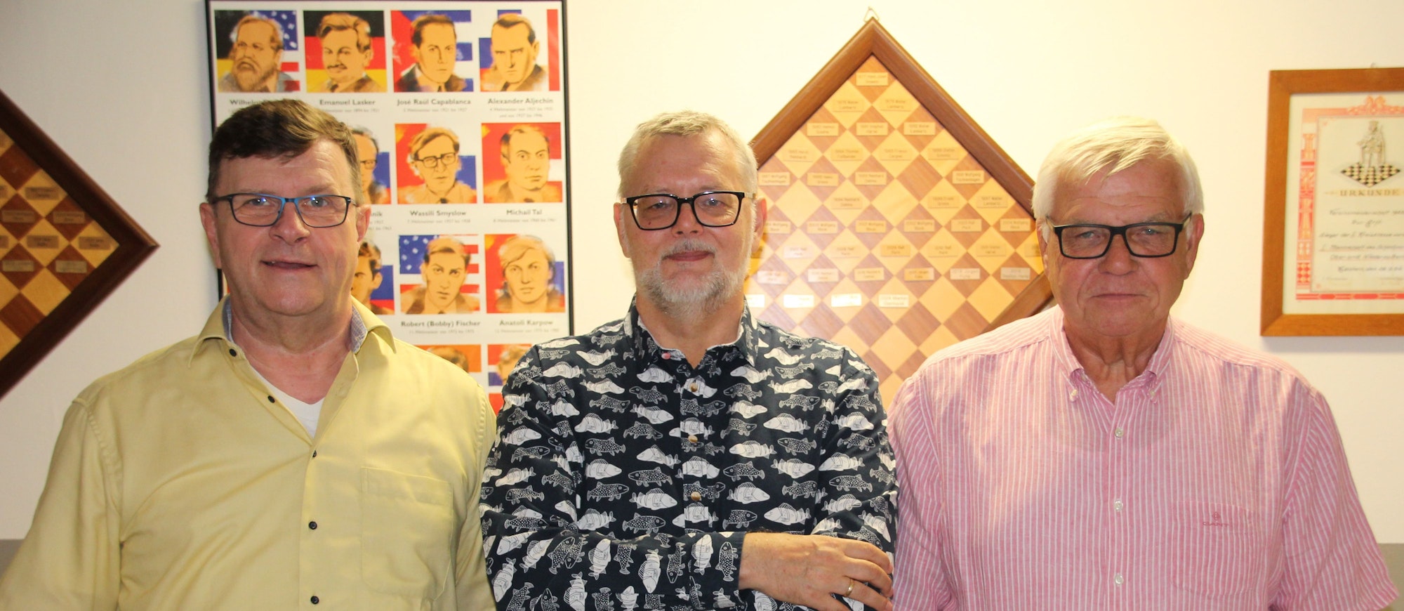 Die drei Männer stehen in einer Reihe, im Hintergrund hängen Spielbretter und Poster als Dekoration an der Wand.