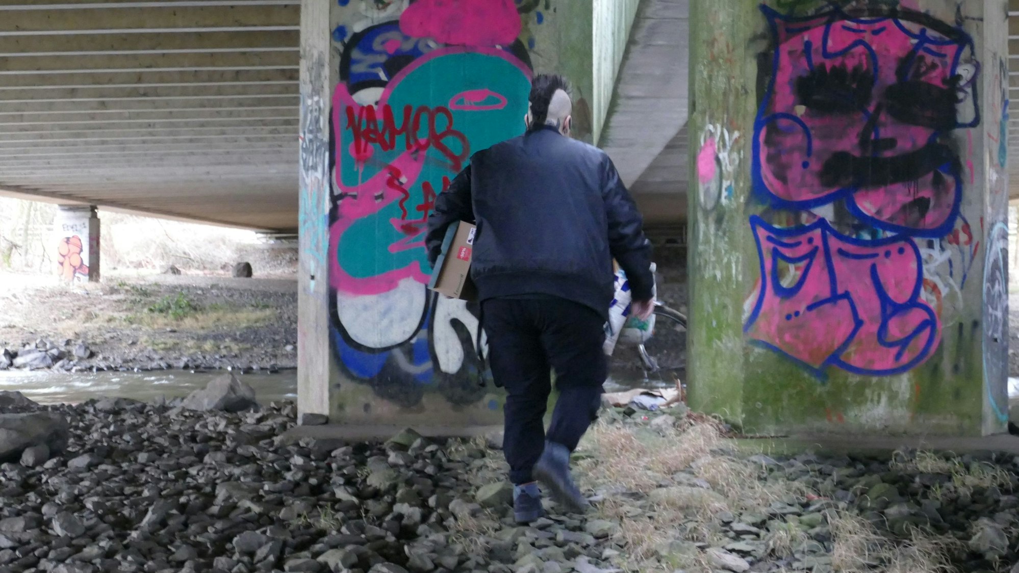 Varressa Lombardi-Boccia trägt einen Schuhkarton unter dem rechten Arm. Die Einstellung zeigt sie von hinten, während sie in Richtung zweier Brückenpfeiler geht.